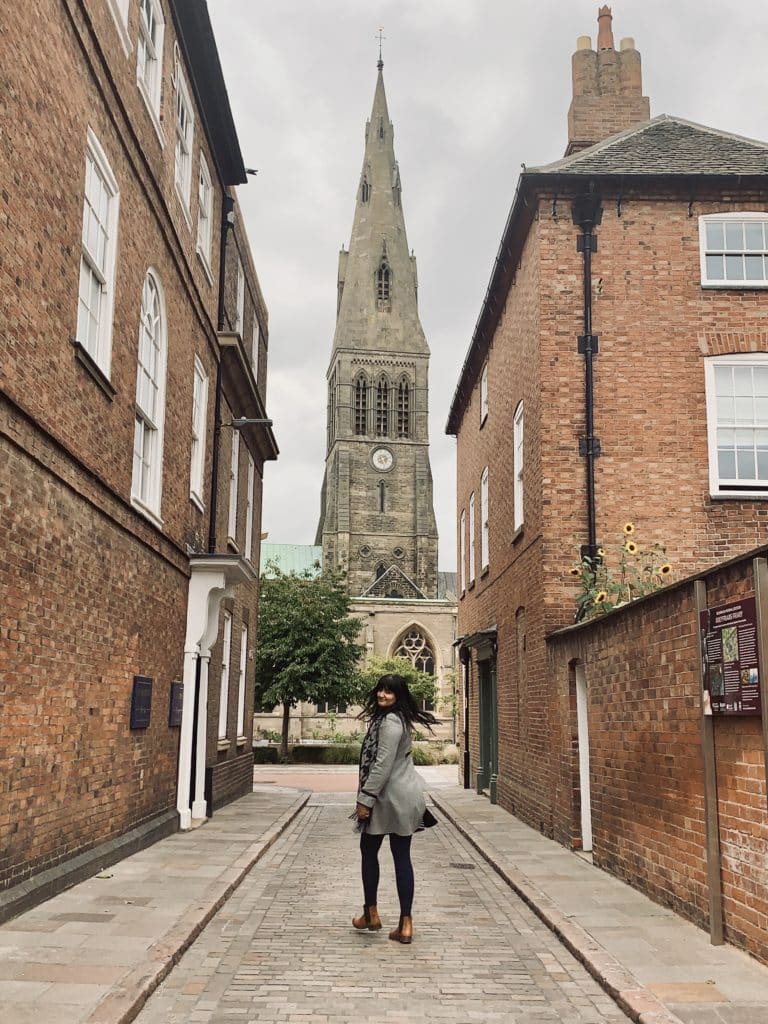 Bejal walking down the street opposite Leicester cathedral wearing a grey coat, brown ankle boot and black tights. The steeple of the cathedral is in the background and Bejal is in mid-twirl to facethe camera. 