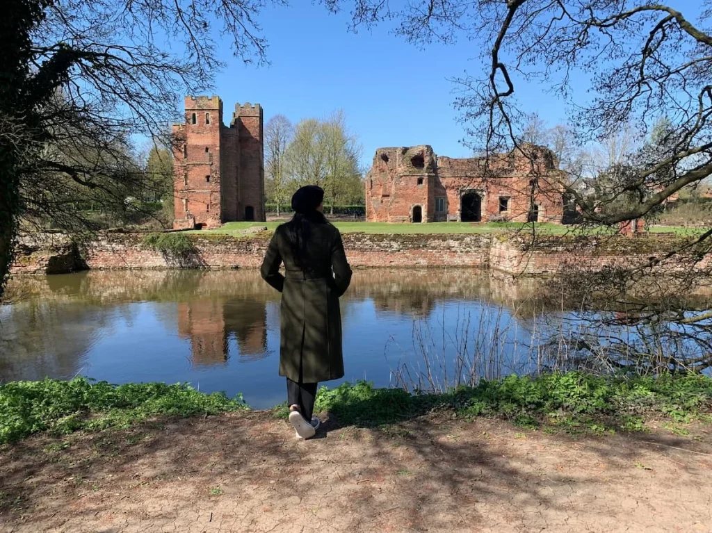 Bejal standing at the back end of Kirby Muxle Castle with a moat around the red brick castle. Bejal is wearing a deep green coat in winter. There are trees and brambles framing the image