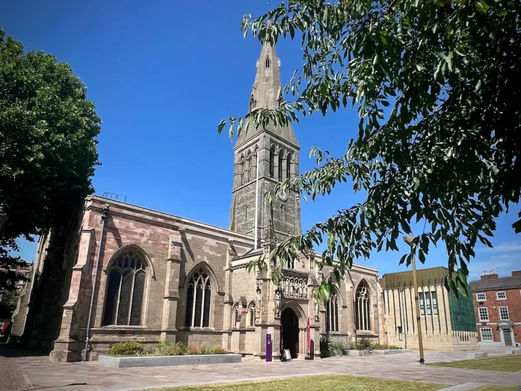 The exterior of Leicester Cathedral with the new Heritage Centre at the end. The image includes the gothic spire and beautiful windows and is framed by trees and foliage