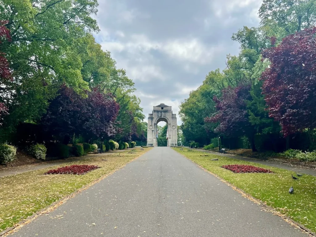 Memorial Arch, Victoria Park with trees lining each side along with flower beds and grass running down both sides of the walkway. The sky is a cloudy blue and the Memorial Arch stands tall in a white and stone colour