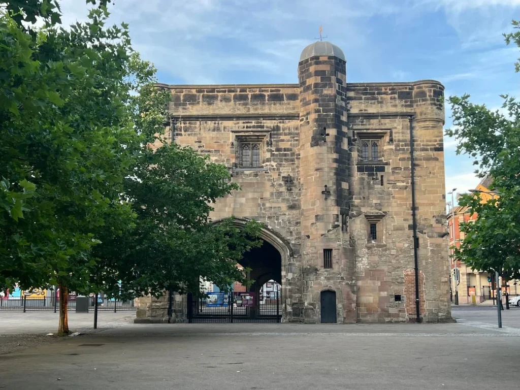 The magazine Gateway light stone exterior with trees hiding the entrance which has a black wrought iron gate across the opening. The road runs down behind this heritage building