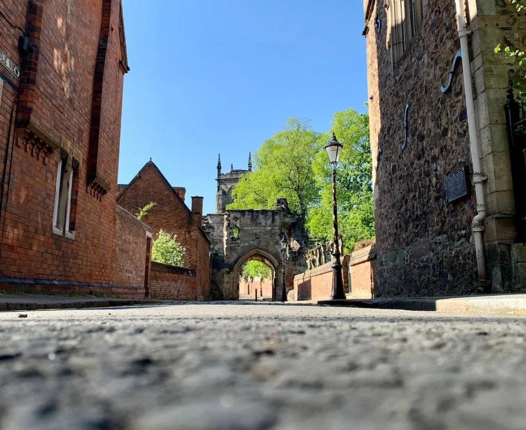 Prince Rupert Gateway in medieval Leicester with red brick buildings on either side. The image has been yaken from the bottom of the pavement, showcasing the grey cobbled stone street. The gateway has trees surrounding it and a bright blue sky.