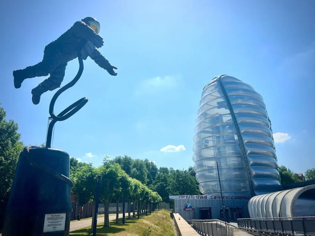 Exterior of the National Space Centre with an astronaut attached to a stand and the Rocket tower in the background and a red sign saying National Space Centre, Leicester