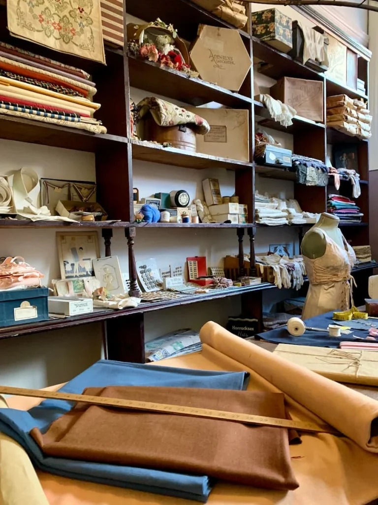 Tailor shop inside Newarke Houses Museum with material, ribbons and cotton on brown shelving. There are rolls of material on the long sewing table in hues of brown