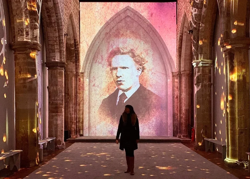 Van Gogh Experience Leicester. Bejal standing in the middle of the immersive experience with a projected image of Van Gogh projected behind her. The light is reflected off the interiors of teh church main hall.