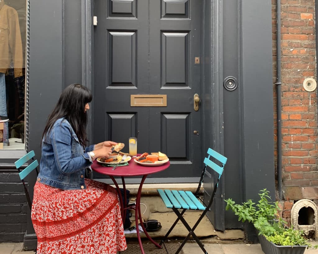 Bejal wearing a red dress and denim jacket having breakfast outside Frank's Bar al in Norwich. There is a big grey door behind Bejal and the table has a pink top and bright lue chairs.
