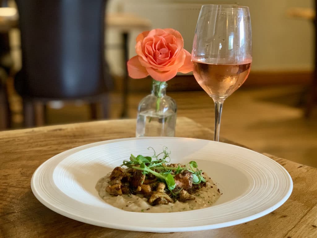 The mains of Norfolk foraged Chanterelles Risotto with crispy shallots, parmesan crisp and shallot on a white plate with a peach rose in a glass vase and a glass of rose in the background, Norfolk
