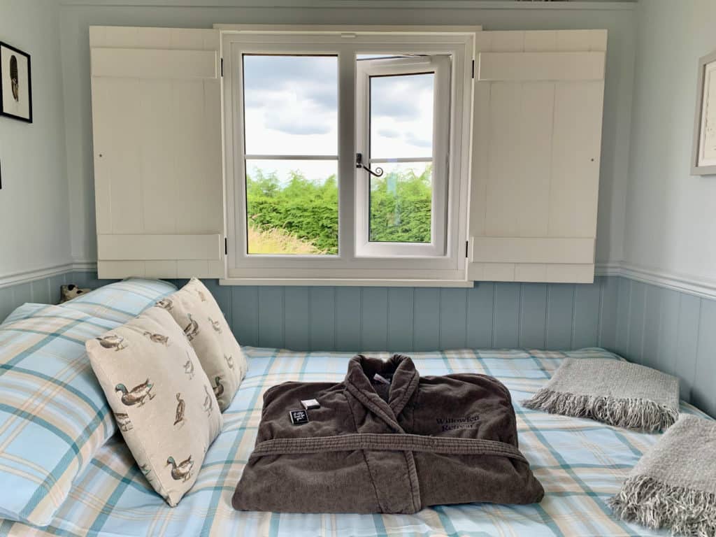 Fluffy dark grey bath robes place son the blue and white checked bed linen with white mallard duck print scatter cushions. There is a window above the bed with views of the countryside at Willowfen retreat, Norfolk