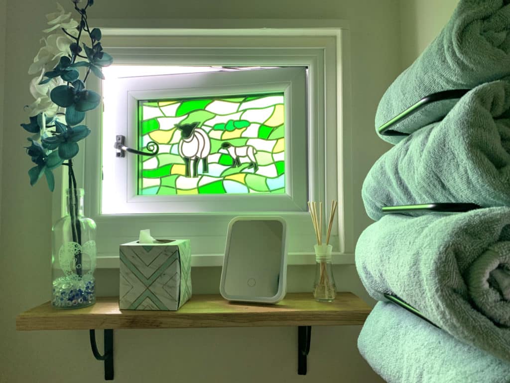 Bathroom Stained green glass window with sheep adn rolling Norfolk hills. There are blue bath towels on the rack with a tissue box, infuser sticks, a mirror and floral display in a vase, Norfolk