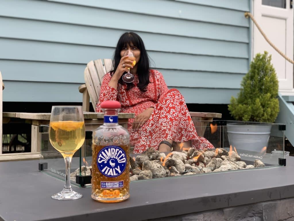 Bejal sitting outside in the garden with in front of teh fire pit. Bejal is wearing a red and white dress and is drinking a glass of Stambecco Italy. There is a potted shrub plant next to Bejal at Willowfen Retreat, Norfolk