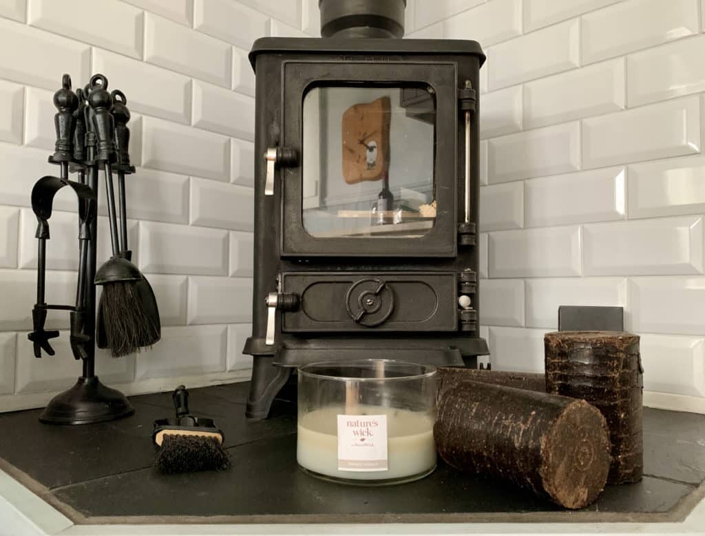 Wood burner with coffee logs in the Shepherd's Hut, Norfolk. The area behind has been tiled with white brick tiles adn there are candles and a chimney brush to the side at Willowfen Retreat, Norfolk