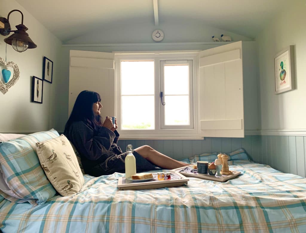 Bejal sitting in a dark grey dressing gown having Breakfast at Willowfen Retreat, Norfolk on the bed holding a cup of tea in had. There are boiled eggs, toast, milk and jam on the bed