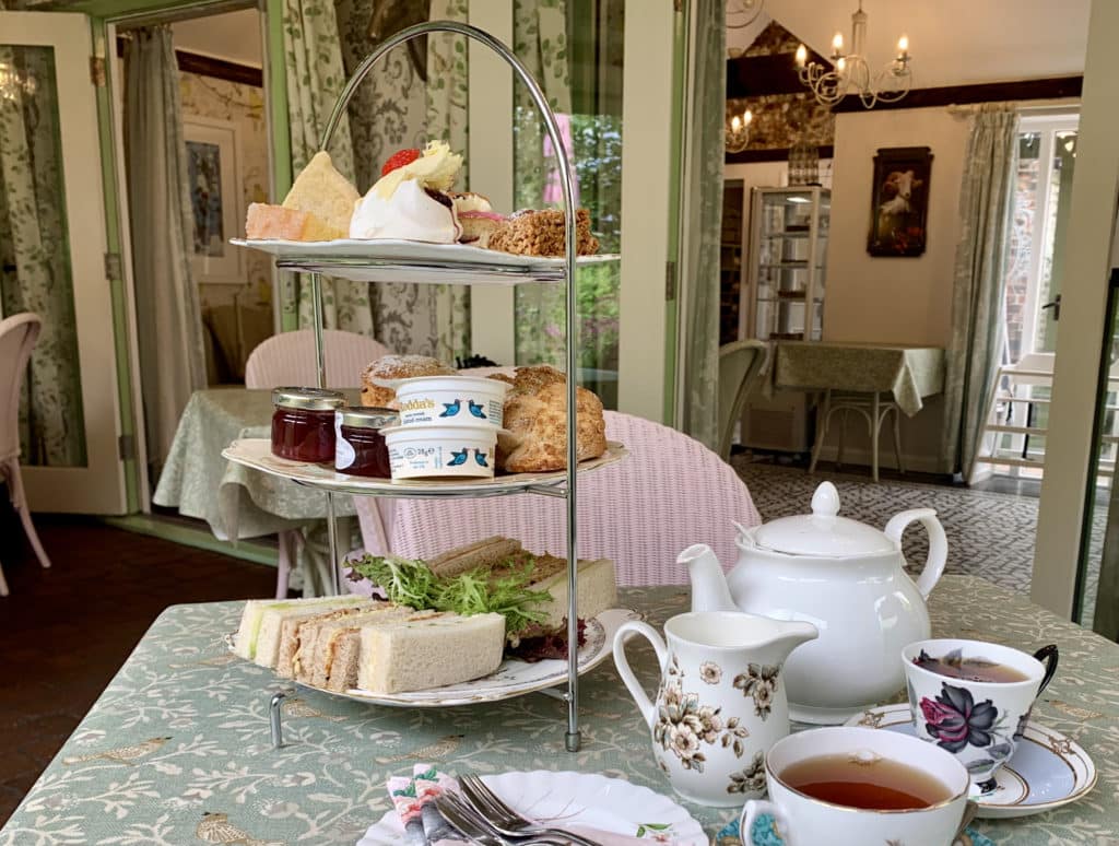 The Folly Tearooms Afternoon Tea cake stand with scones, sandwiches, clotted cream, jam, pastries and tea cups full of tea. In teh background in the sage green floral wallpaper decor,Norfolk