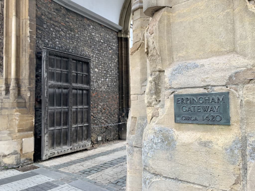 The grey sign for Erpingham Gateway Circa 1420 on light beige stonework with a bronw gate in the background