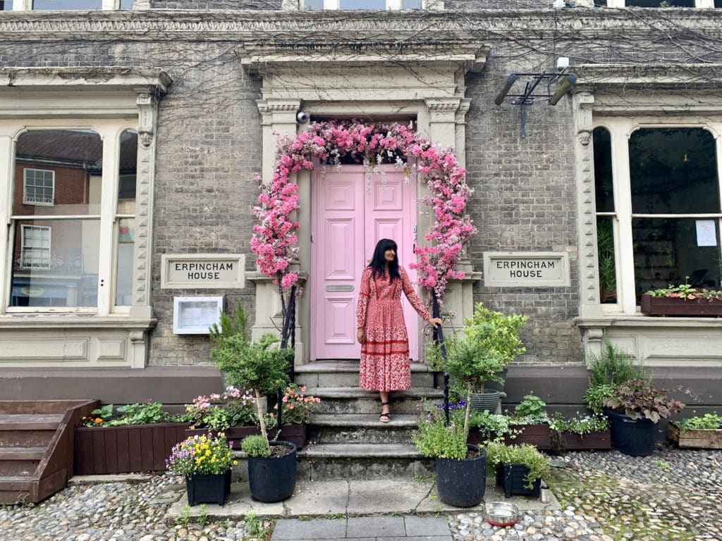 Bejal wearing a red and white dress standing outside a pink door adn floral archway at Erpingham House i Tombland. There are potted plants at the entrance of the pink door