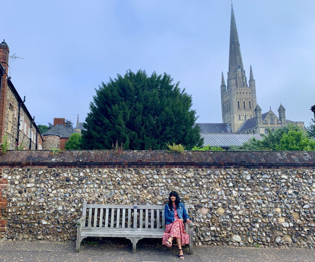 Very best of Norwich: Things to do in 2 Days: Bejal sitting on a bench wearing a red dress and denim jacket with Norwich cathedral behind her, atree and bright blue skies.