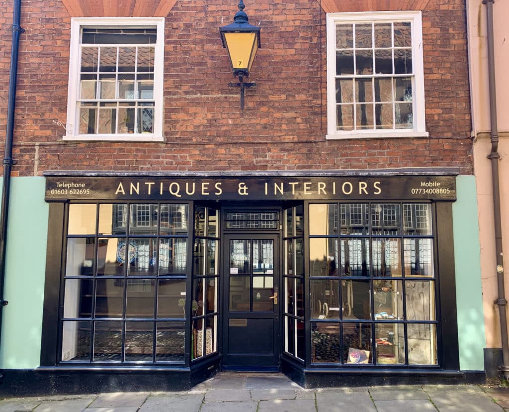 The exterior of the Antiques & Interiors Shop on Elm Hill in Norwich. The outside is black with a double-faced front and pale green sides with a brick portion at the top.