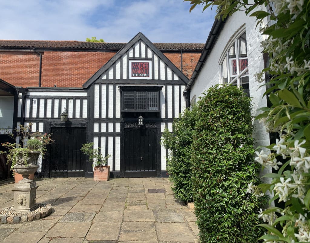 The Tudor style exterior of the Maddermarket Theatre, Norwich. There is foliage and green tees to the left hand side and a paved grey floor outside.