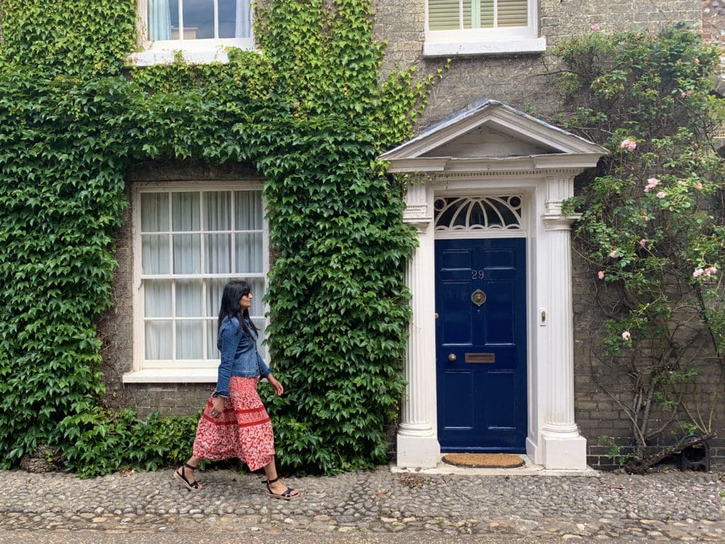 Bejal wearing a red and white dress and doing a stride by outside the Cathedral Quarter houses. The house has a navy blue Regency style door adn there is ivy growing on the walls with a rose bush to the right.