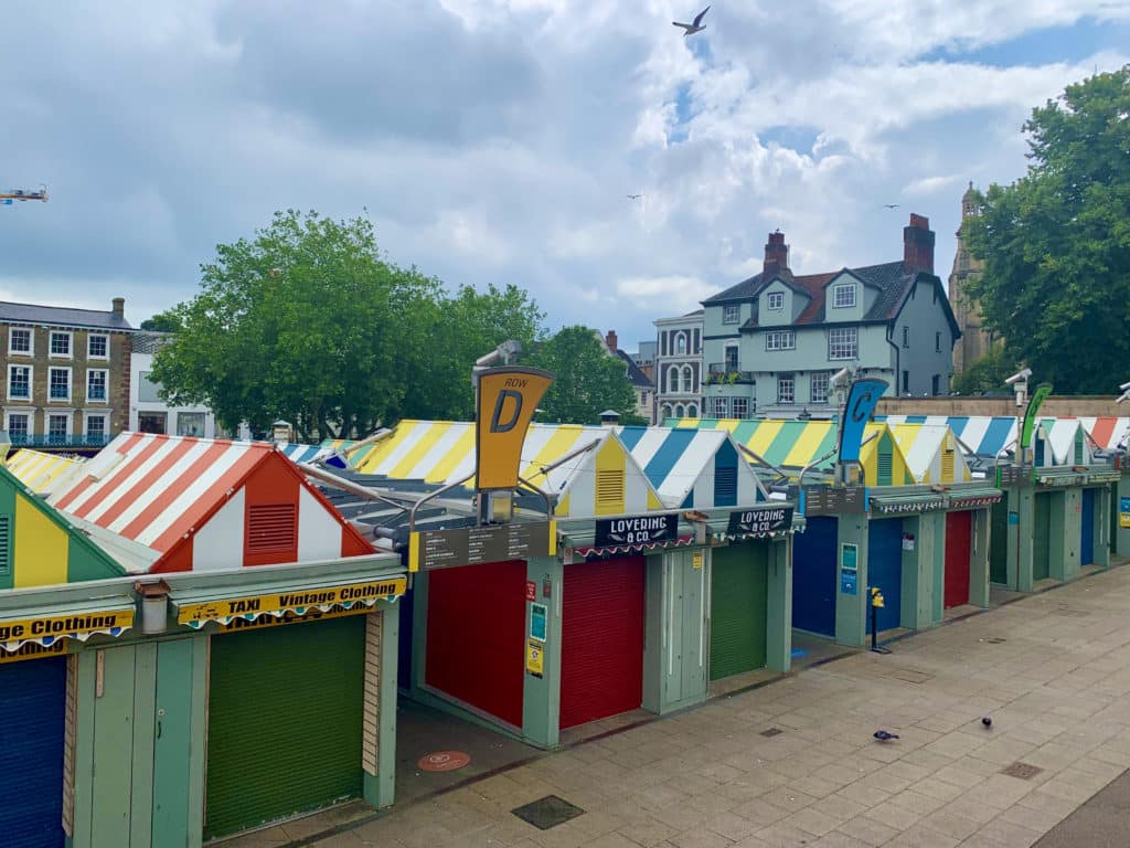 A row of colourful market stalls at Norwich Market. Stand D is in the image with striped red, blue and yellow surrounding stalls.