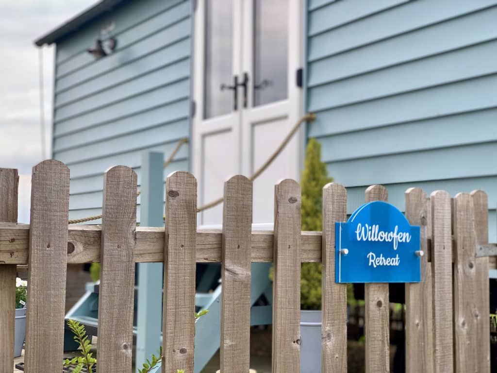 Willowfen Retreat Norfolk, Sheherd's Hut from the exterior. It is painted in duck-egg blue with 2 wooden chairs outside and a electric fire with potted plants and foliage surrounding the hut. There is a blue Willowfen Retreat plaque in white writing on the wooden entrance fence