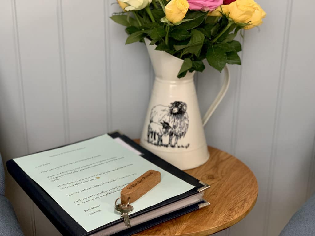 A cream coloured vase, with black sheep outline. The vase is filled with yellow and pink roses and there is a welcome manual and keys to the side of the vase. They are both on a wooden circular side table and the walls are painted a light blue with stripes at Willowfen Retreat, Norfolk