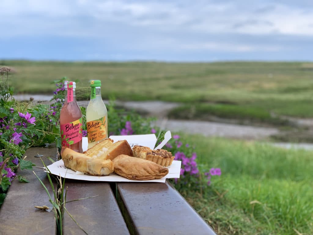 A picnic with pastries, cakes and pasties, Belvoir lemonade from Thornham deli. The picnic has been set up at Thornham Marshes on a bench with the sea in the background and grasslands surrounding, Norfolk