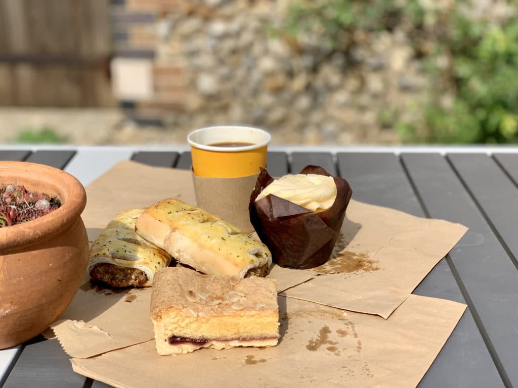 The Art Cafe, Cley, Norfolk. There are vegan sausage rolls and a cupcake on brown paper bags on the table with a coffee in a paper cup in the background.