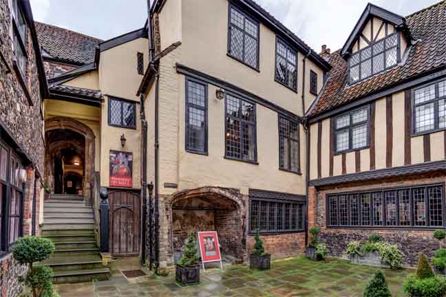 Stranger's Hall Museum exterior in Norwich with its cream and brown medieval architecture and grey skies. The courtyard outside has shrubs nad paving stones.
