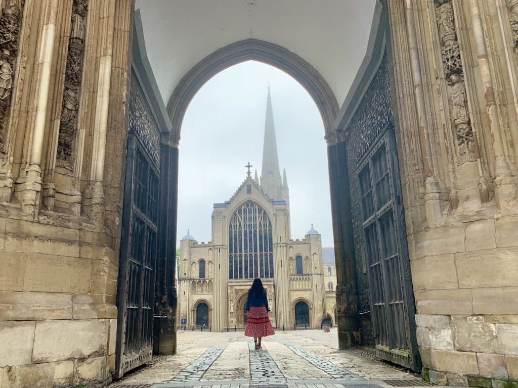 Bejal wearing a red dress and denim jacket walking through Norwich Cathedral West Gate with teh cathedral as a backdrop