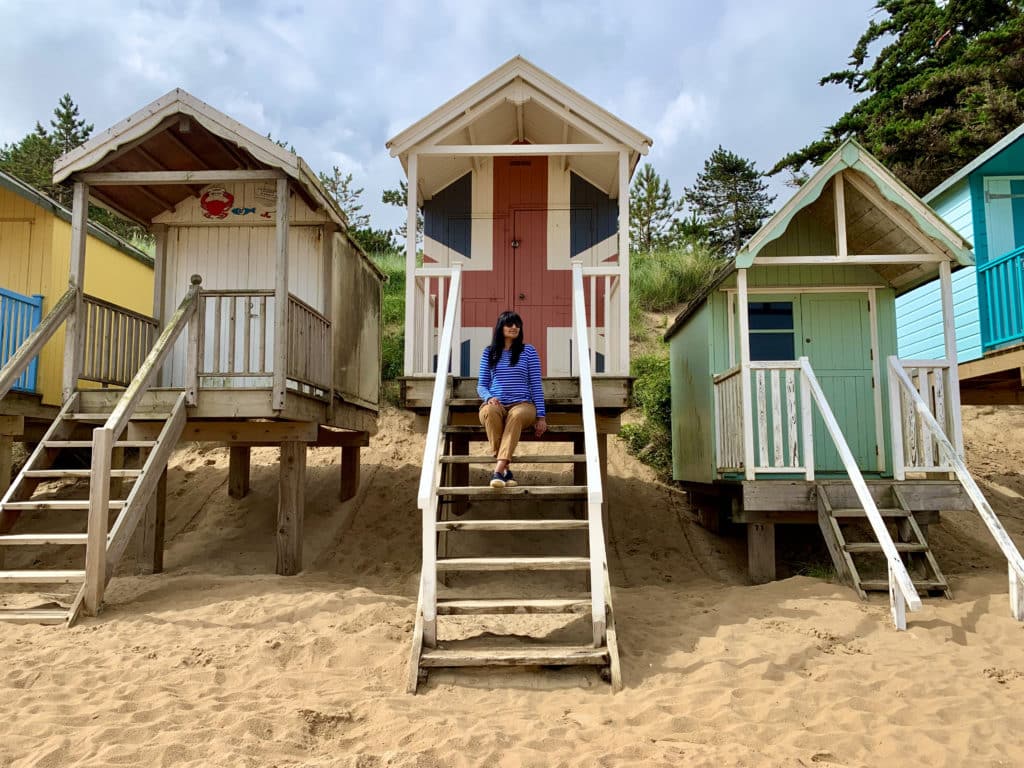 Bejal wearing a blue and white Breton sitting on the steps of a beauch hut painted in the union jack at Wells-next-the-Sea beach with huts at either side