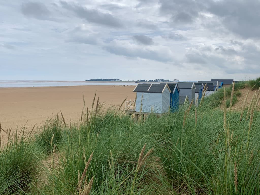 Wells next the Sea beach with its expasive sand and different hsades of blue beach huts to the right. The sea can be seen in the distance with the tide going out. The huts look as if they are emerging from the long grass near the dunes.