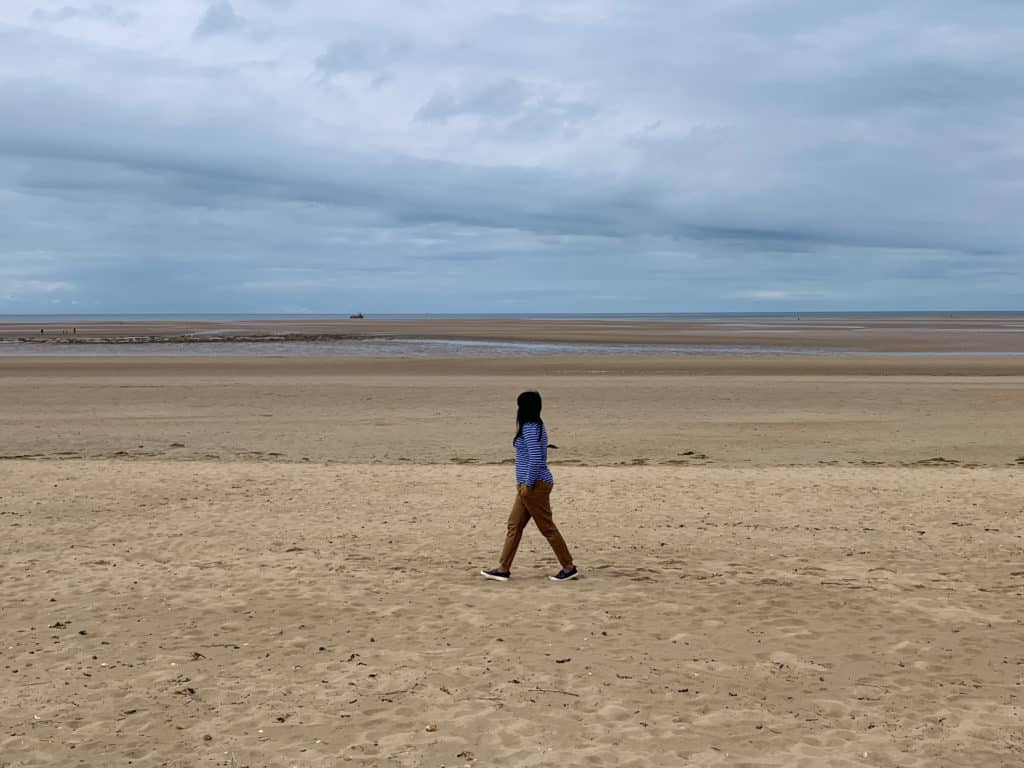 Bejal wearing a blue and white striped Breton with beige chinos walking down Brancaster Beach, Norfolk on the sand. The sky is a cloudy blue almost turning into a storm