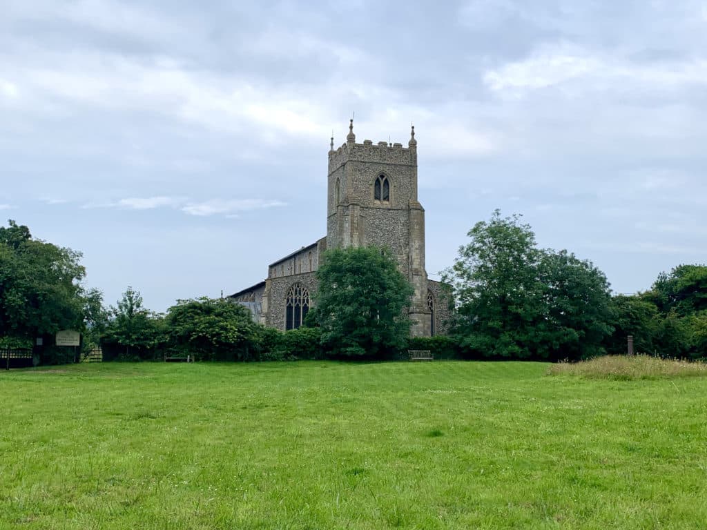 Wiveton Church, Norfolk surrounded by trees and bushes and a lawn in the front. The sky is blue filled with clouds