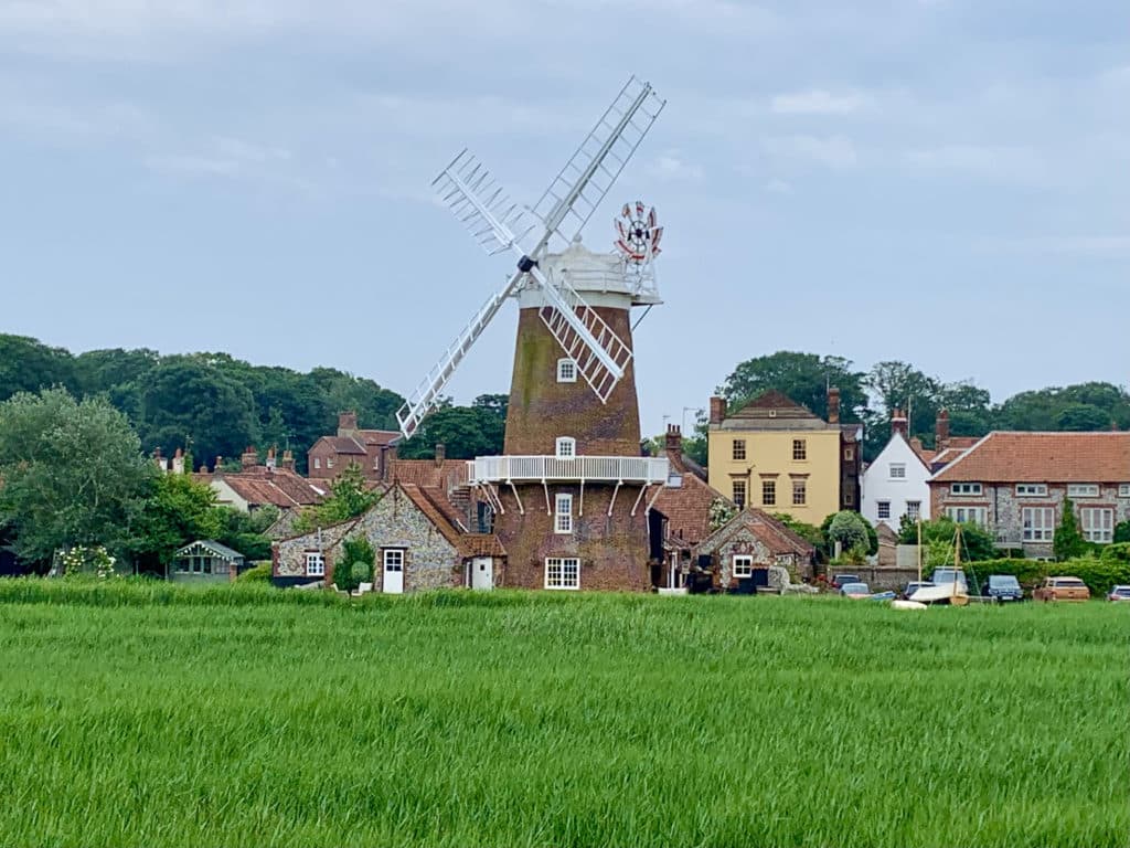 Cley Windmill, Norfolk in Cley-next-the-Sea. Photograph taken from the distance with a stretch of green grass in front of the windmill which has white sails and a grown stone exterior. The sky looks pretty cloudy in the background
