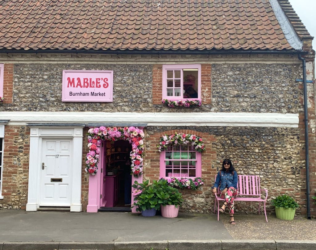 Bejal wearing pink floral printed trousers and a denim jacket sitting on the pink bench outside Mables, Burnham Market, Norfolk. The pink door and window are dressed in faux flowers.