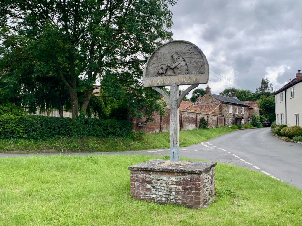 Bawburgh, Norfolk village sign set in bricks and stone in the centre of teh village with traditional houses in the background the village green surrounding