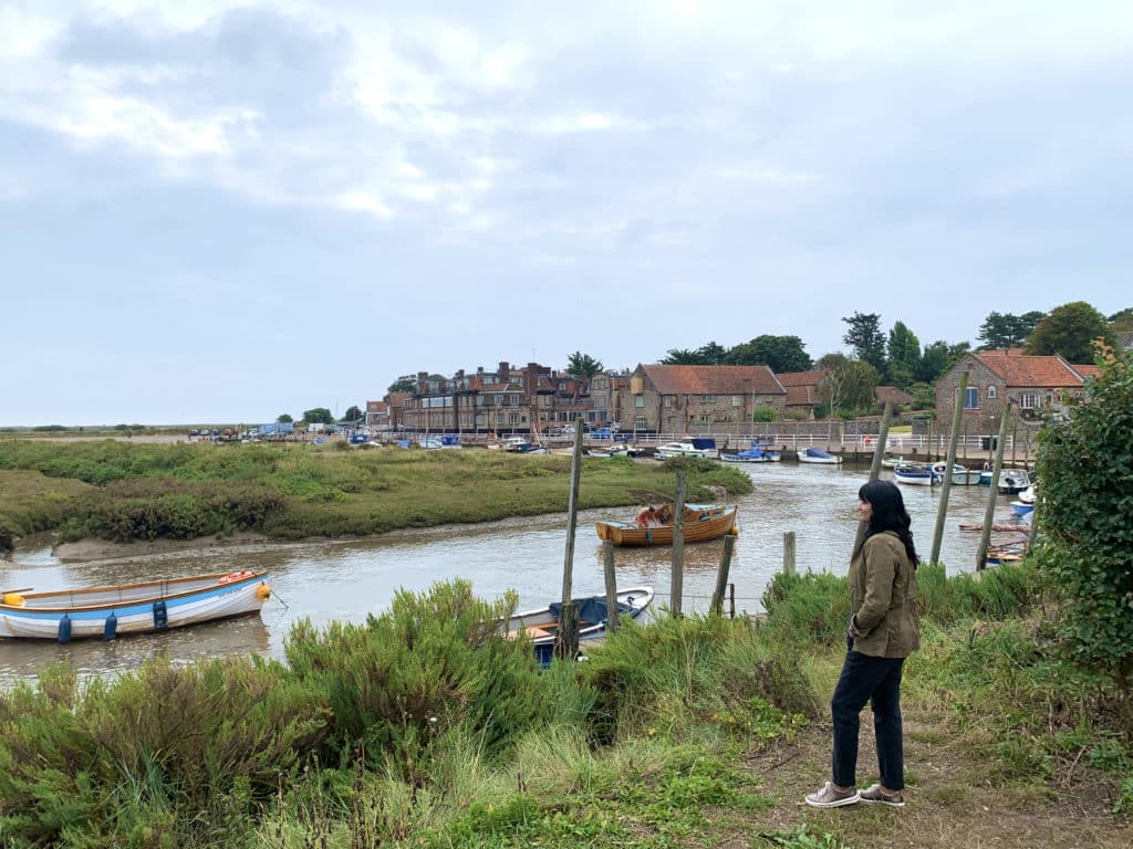 Bejal standing looking out to Blakeney Harbour in Norfolk. There are fishing boats and the Blakeney Hotel in the background.