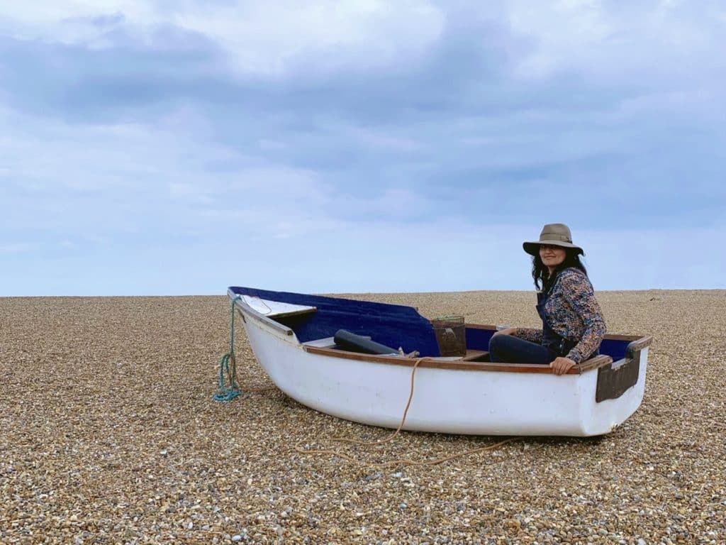 The best of Norfolk: Bejal sitting in an old fishing boat on Cley beach which is covered in Shingles. The sky is a pale blue and slightly cloudy. Bejal is wearing a floral top with black dungarees