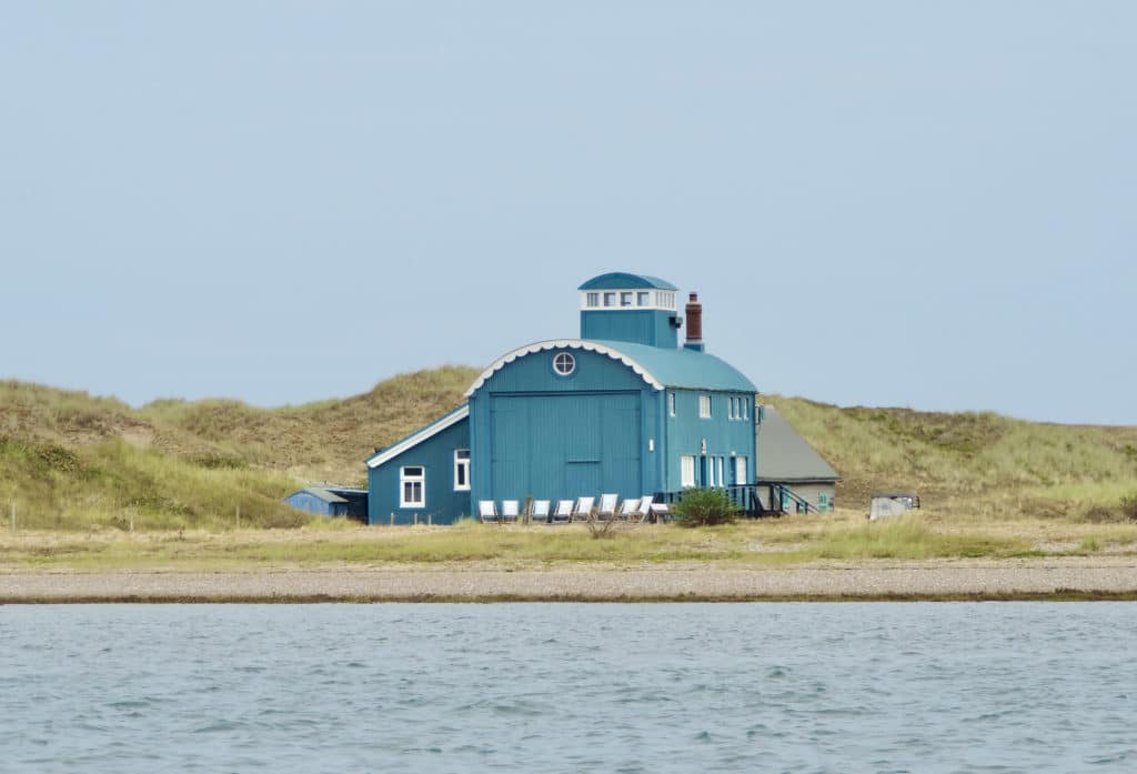 National Trust Morston Quay building from a boat. The building is on an island in Norfolk and is painted in a duck egg blue adn white with deckchairs outside. There are grasslands surrounding the building.
