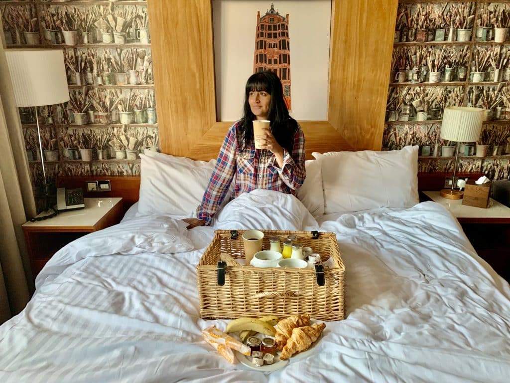 Bejal sitting in bed having breakfast from the breakfast hamper at Abode Hotel, Glasgow