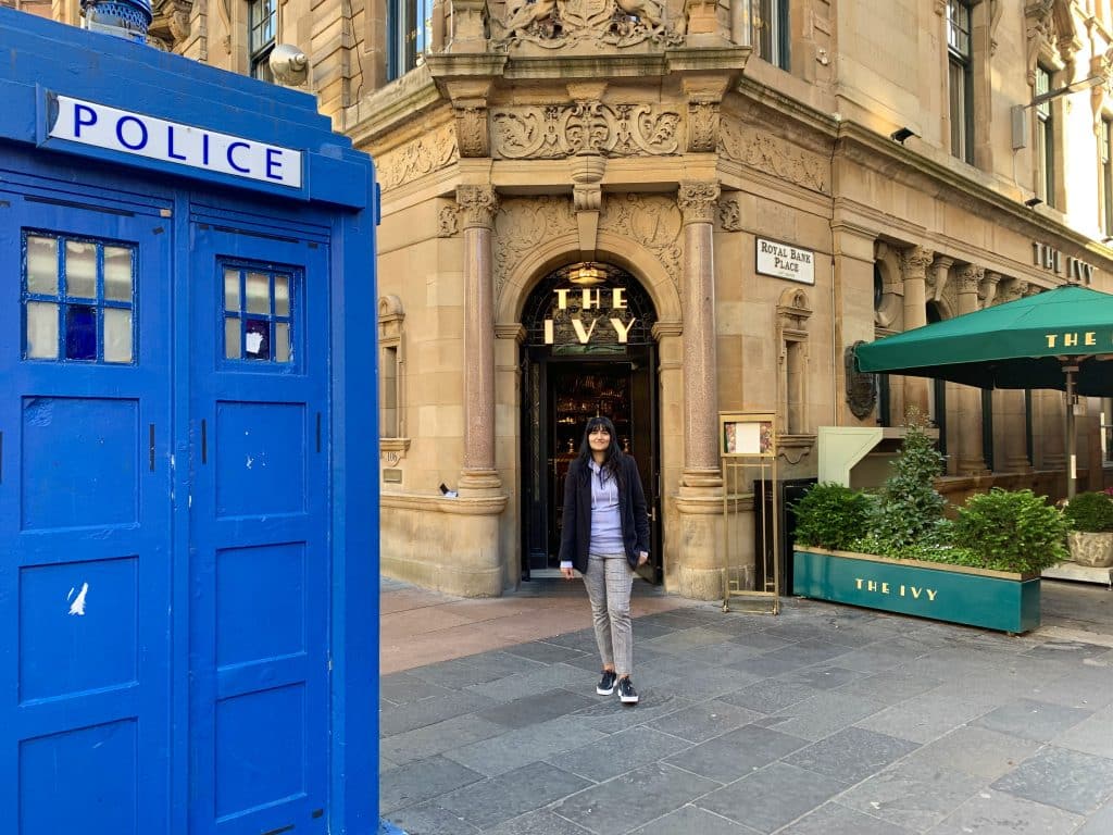 48 Hours in Glasgow: Glasgow Streets with Bejal standing in the middle wearing grey trousers, a hoody and a black coat. A bright blue police box is to the left.