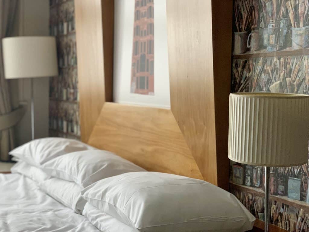 ABode Hotel Glasgow Bedroom with white pillows and bookcase wallpaper background with 2 bedside lamps