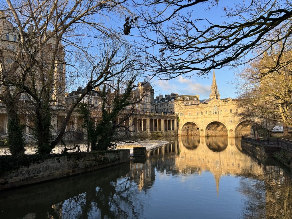 Weekend Guide to Bath: Bath Pulteney Bridge and surrounding historic buildings with the River Avon abd tree branches framing the photo