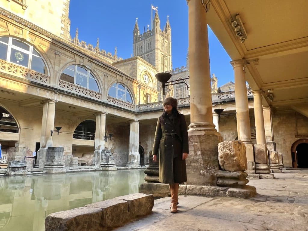 Bejal wearing a khaki wool military coat, faux fur hat and tan boots standing in the Roman Baths in Bath. Bath Abbey is in the background against a bright blue sky