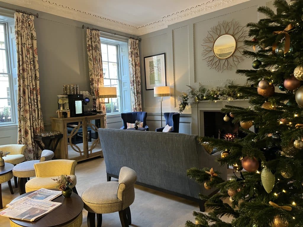 The lounge at Queensberry Hotel, Bath which has a huge dark grey  sofa and light grey chairs and a fireplace. There is a coordinating Christmas tree to the side as we visited at Christmas times