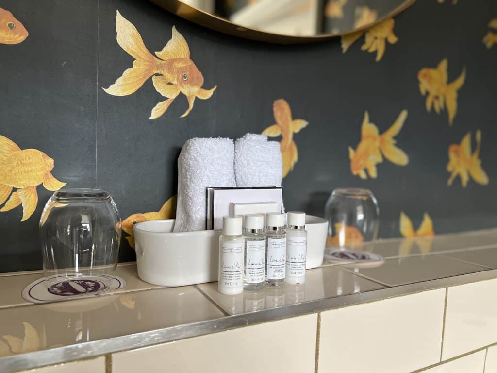 Bathroom Toiletries at The Queensberry by The White stuff on the bathroom shelf with mustard goldfish wallpaper on a dark grey background