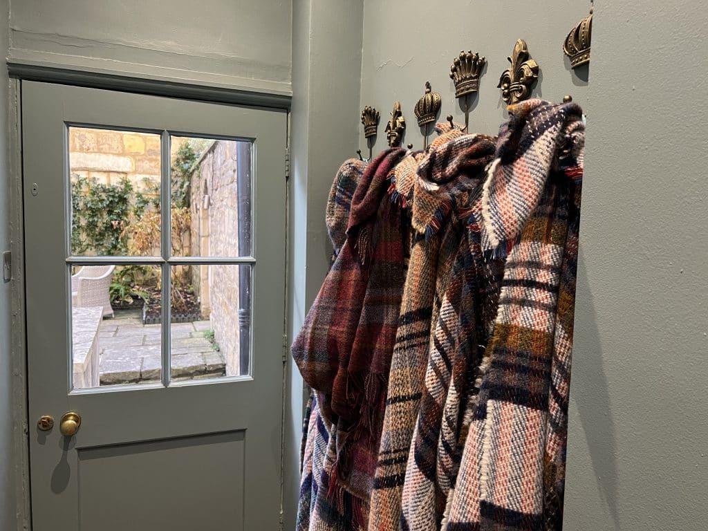 The Queensberry Hotel, Blankets Blankets for the terrace in the hallway leading to the outdoor space. They are checked and hug on gold hooks on the wall, The sage green walls and door lead outside, with a glimpse of the foliage in the garden