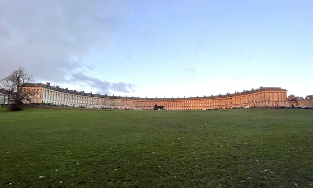 A panoramic shot of The Royal Crescent at golden hour in Bath. There is a green lawn right in front of it