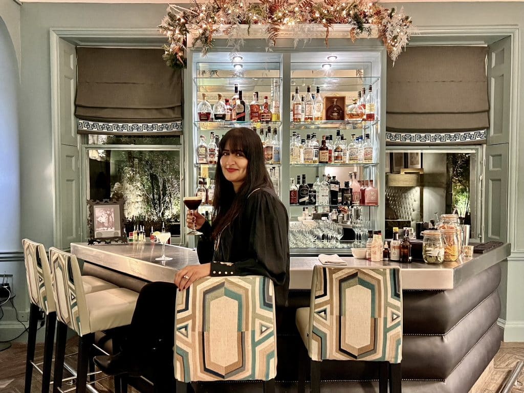 Bejal wearing a black shirt sitting at the Queensberry Hotel, Bath bar holding an Espresso Martini in her hand. The chairs have a pastel design on then and bar is an assymetrical shape